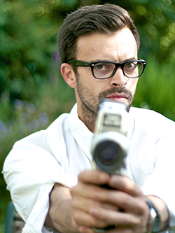 A young man in dark-rimmed glasses brandishing a video camera as if it was a gun