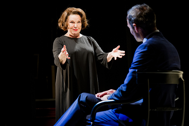 A middle-aged woman in black with vivid red lipstick explains something to a suited man sitting in a chair.