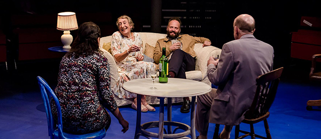Two men and two women drink wine around a table on a stage set