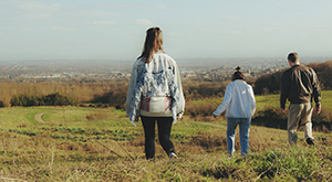 Two young women and a man in 80s clothes stand in a field with their backs to us, looking towards a town on the horizon